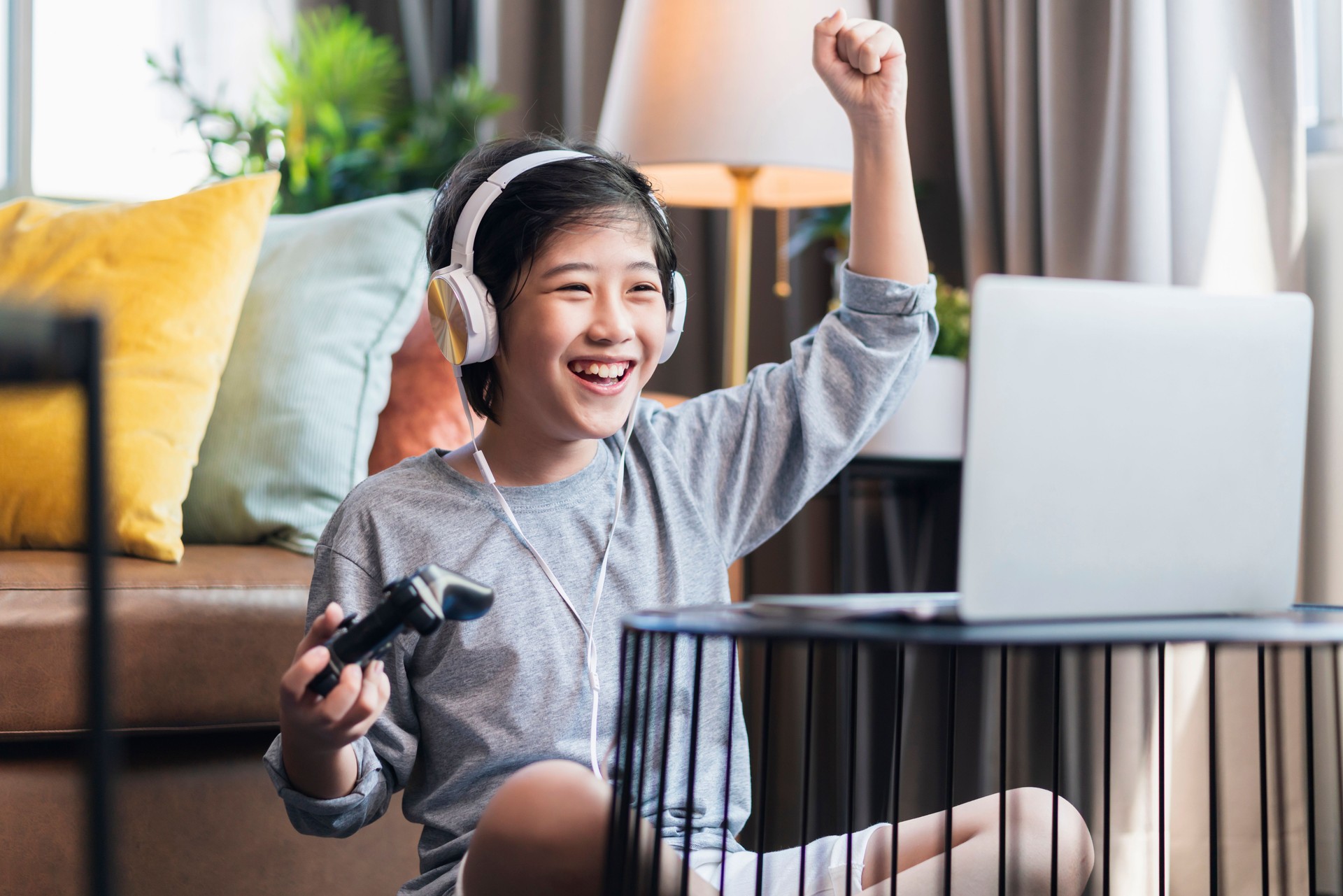 chilling weekend holiday with esport game online,asian child boy wear headphone hand playing laptop game online with exited and fun sitting  on floor at living room at home, son is playing alone with mobile equipment.